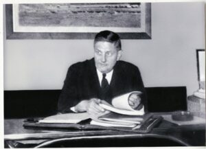 white man in dark suit at desk with papers