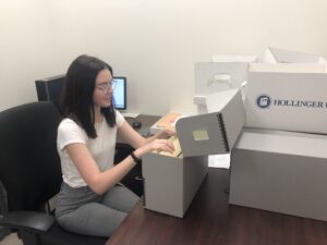young white woman with long dark hair and glasses sitting in office chair at desk with archival boxes