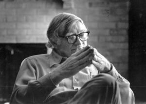 older man with combed over hair and glasses sitting with his hands out and fingertips touching