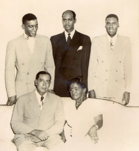 an older Black man and Black woman seated on white couch with three younger Black men standing behind them in suits