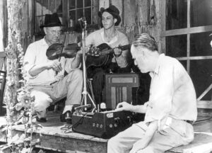 white man with recording machine on porch with older white man with fiddle and younger white man with guitar