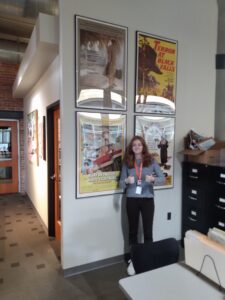 white young woman with long brown hear wearing a sweater and dark pants standing in front of movie posters on a white wall