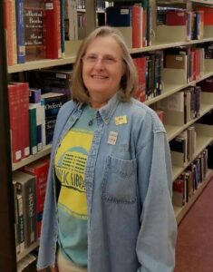 woman with blond hair and glasses in blue tee shirt with blue long sleeved shirt over it next to library stacks