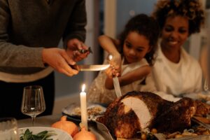 young Black girl helping an adult carve a turkey on a table with a smiling Black woman in the background