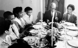 two older African American people and several teenage African Americans sit around a table with plates and candles