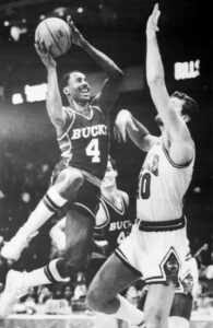 one African American man leaps holding a basketball while a white man tries to block the shot with his arm