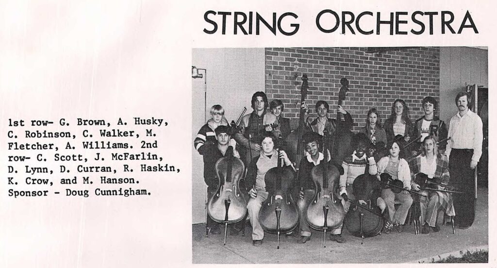 group of students with orchestra instruments posing in front of brick wall with white male teacher at the far right