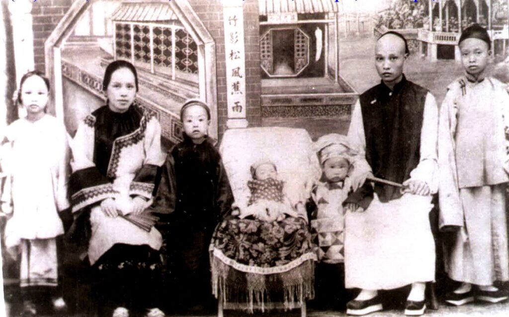 a Chinese family of seven lined up for a photo with a baby in a bed in the center next to a toddler in a hat