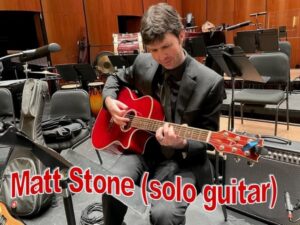 photograph of a man in a music room wearing a black suit holding an acoustic guitar with the words 'Matt Stone (solo guitar)'