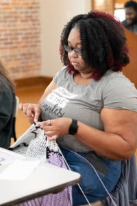 black woman with gray shirt and glasses knitting something purple and white