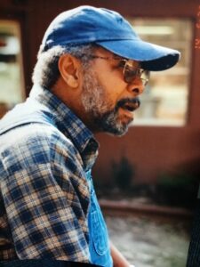 African American man with beard, mustache, glasses, blue ball cap, wearing plaid shirt and overalls