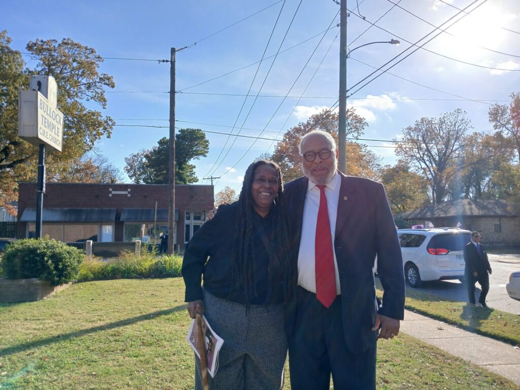 African American woman with long dreadlocks and African American older man in red tie stand next to each other with his hand around her shoulder with a sign reading Bullock Temple behind them