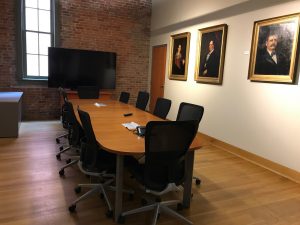 meeting room with long wooden table wheeled chairs and large TV on wood floor with brick wall and plaster wall with three portraits