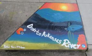 Fish swimming in river under sunset with "Drain to Arkansas River" text on sidewalk storm drain