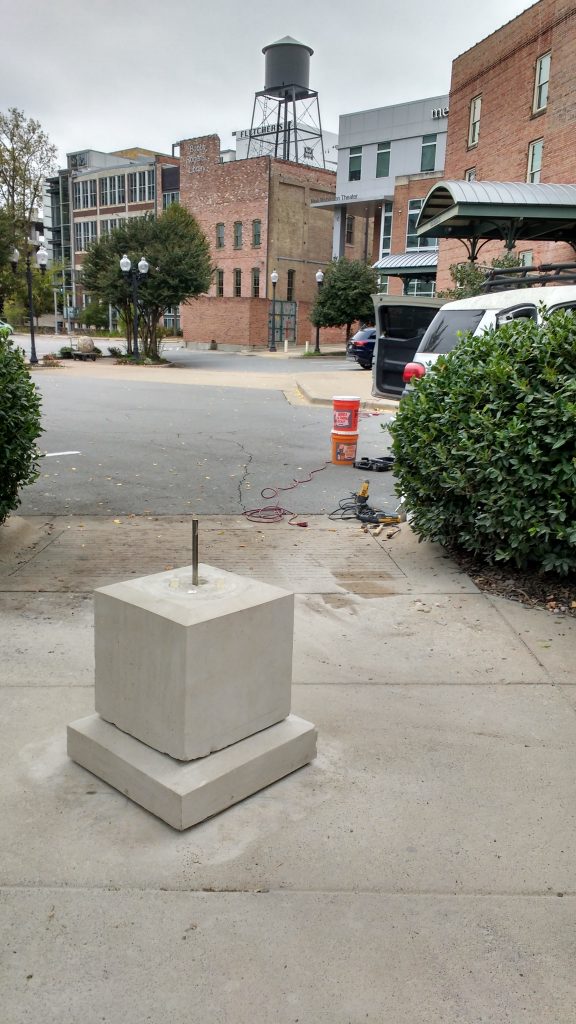 Stone pedestal on sidewalk with multistory buildings and parking lot in the background