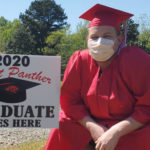 Young white man in red cap and gown and face mask kneeling next to "2020 Cabot Panther Graduate Lives Here" sign