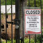 "The Old Mill is closed to minimize the spread of covid-19" sign on locked gate with mill building in the background