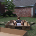 White boy sitting in carboard lid with paddles on his front yard