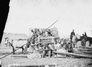 horses attached to a partly covered wagon with black people in and around it and uniformed white men in the background