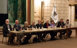 group of white men and women with most wearing suit jackets sitting at a long table with flag in background