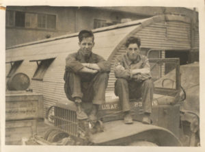 two young men in military pants and shirts sitting with arms crossed on truck in front of corrugated building