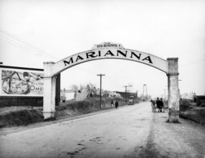 Arch reading "the city beautiful Marianna" over dirt road with people, cars, and a wagon
