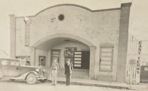 one-story building with arched entrance with car and two men in front