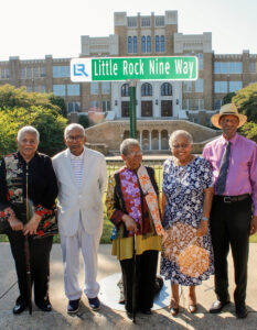 Five African Americans standing beneath a sign "Little Rock Nine Way" in front of a large brick school building