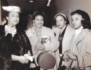 Older African American woman with fur coat and hat standing with two younger and one older African American women in coats