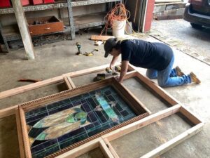 woman wearing ball cap down on knees working to put a stained glass window into a wood frame