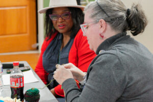 black woman in hat glasses and red sweater watching white woman with glasses and ponytail working with yarn