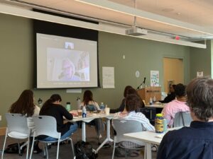 view of the backs of adults sitting in a classroom looking at a white man on a projected computer screen
