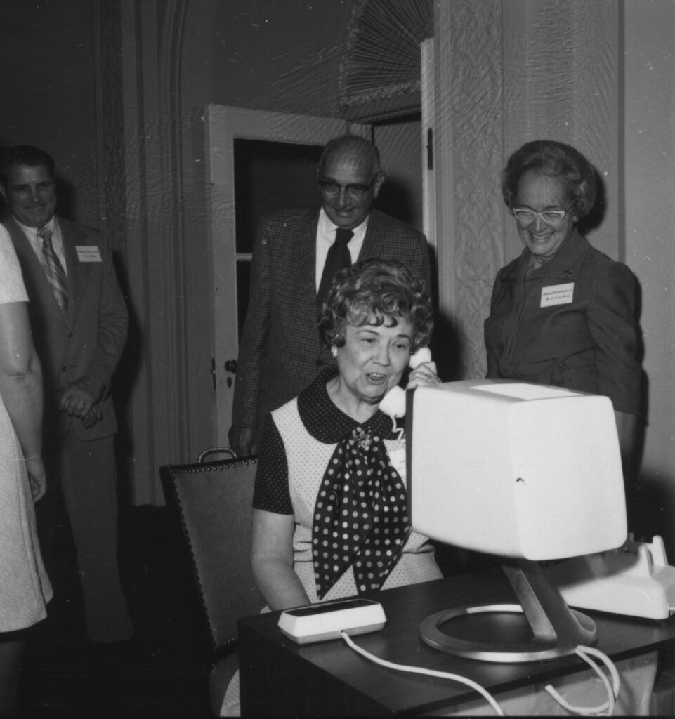 white woman with polka dot scarf sitting in front of a large monitor with a phone receiver in her hand while others look on