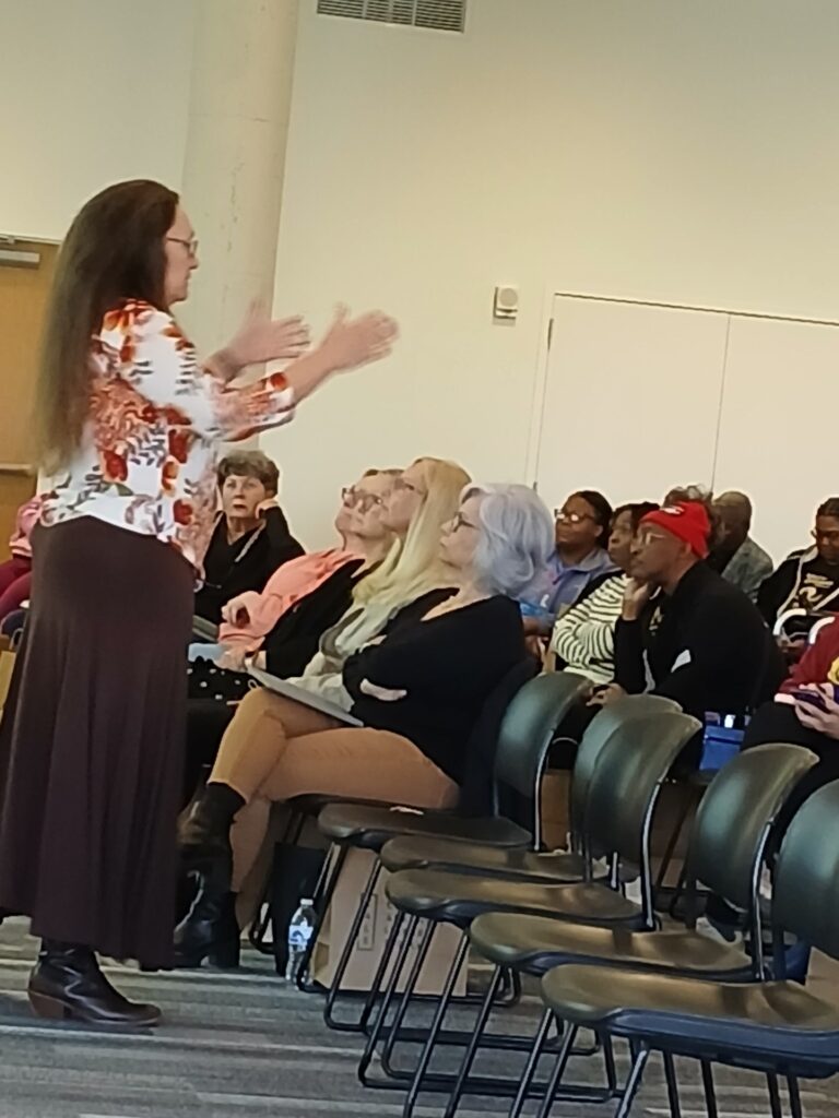 white woman with long brown hair and brown skirt standing in front of seated audience members