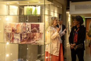 white woman with light hair and red skirt talks with black woman with glasses and black blazer in front of glass case containing open books standing up