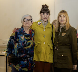 older woman with colorful blouse and black rimmed glasses, middle age woman with bangs and yellow coat, and middle age woman with shoulder length blond hair and green military style jacket