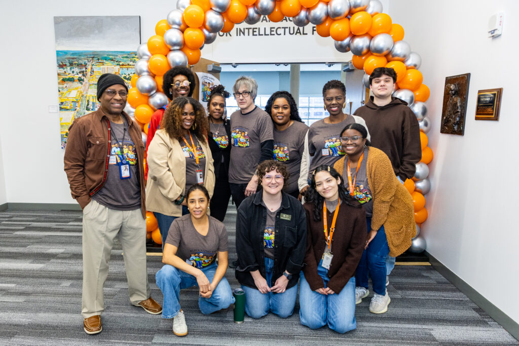 a group of people wearing matching gray decorated t-shirts posing in front of an archway of orange and silver baloons