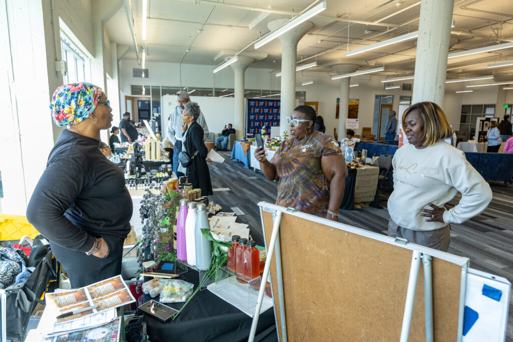 Black woman in black shirt and colorful headwrap standing behind table with objects on it while two Black women observe