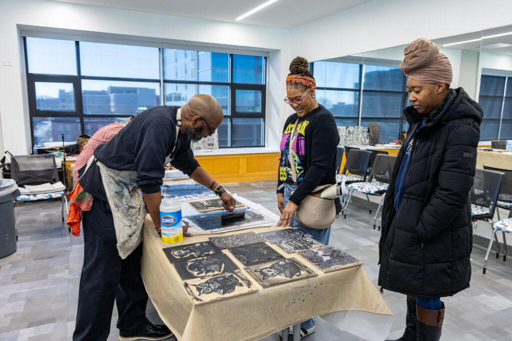 bald Black man in dark pants and shirt leaning over table with black ink and printmaking supplies with two Black women observing