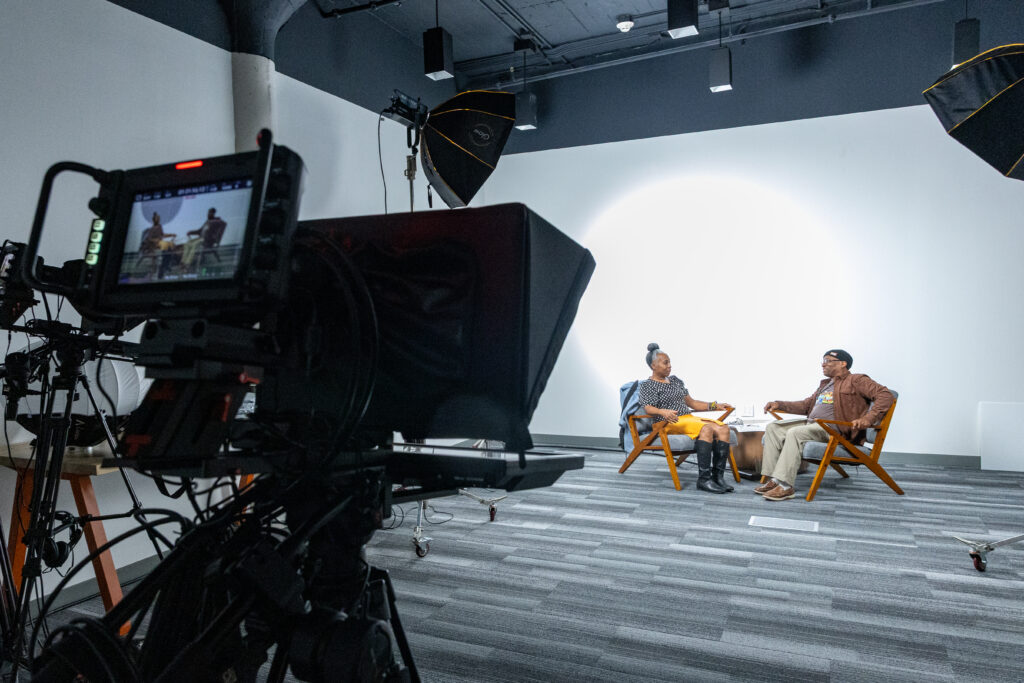 a video camera in the foreground with a woman and man sitting in chairs at the back of the frame