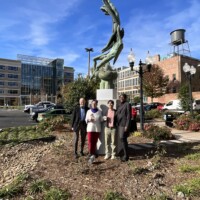 Jim Wohlleb, Mary Wohlleb (granddaughter of Raymond Rebsamen), Ariana Remmel (Rebsamen’s great-grandchild), and CALS Board President Karama Neal four people stading in front of tall statue in front of buildings and blue sky